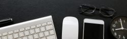 Tastatur, Maus, Smartphine, Uhr und Lesebrille liegen auf einem schwarzen Schreibtisch