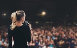 Frau mit Mikrofon vor Publikum, Thema Rhetorik